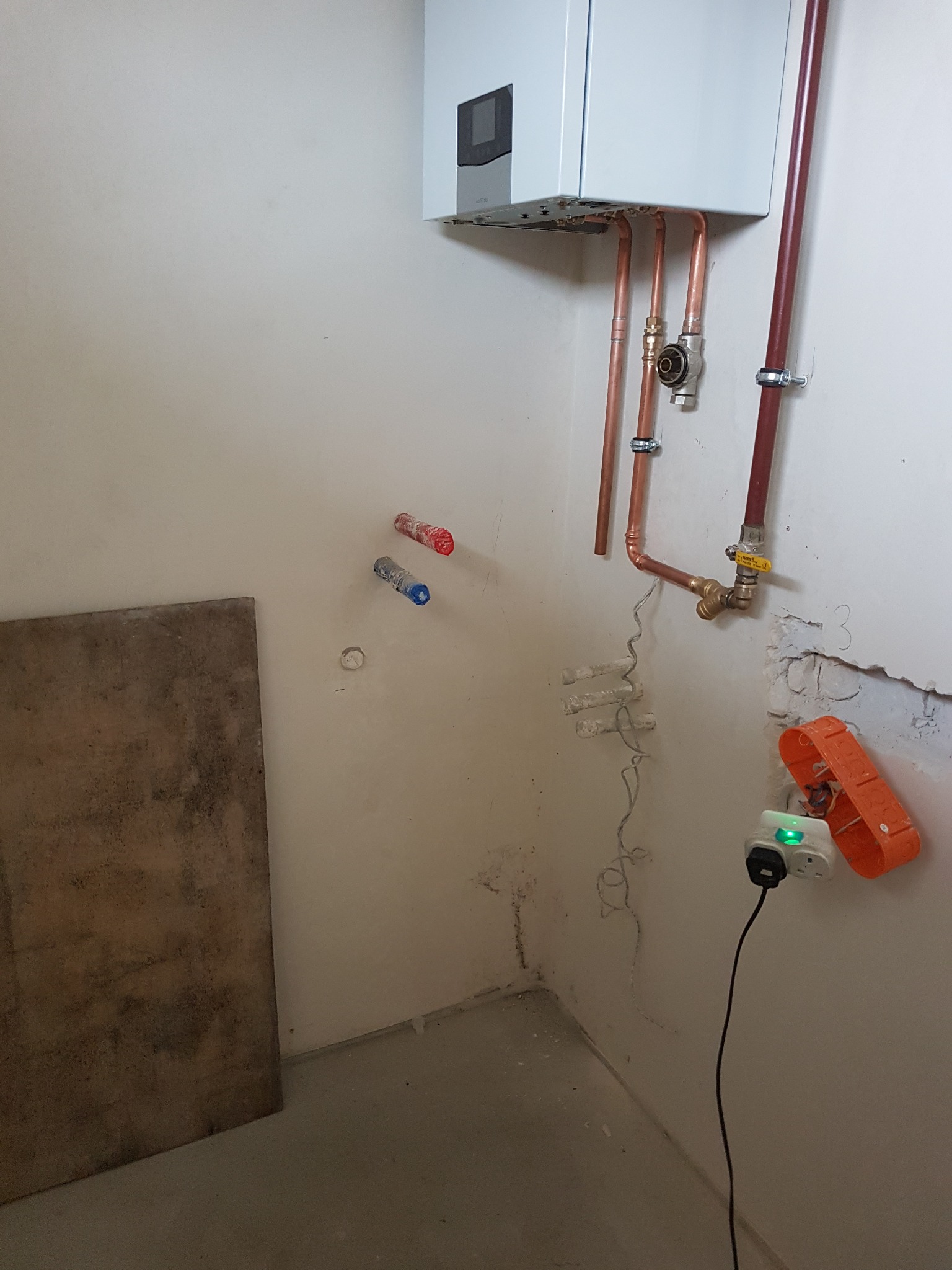 Biały piec gazowy zamontowany na ścianie z widocznymi miedzianymi rurami instalacji. Fragment ściany z uszkodzeniami i wystającymi przewodami elektrycznymi.