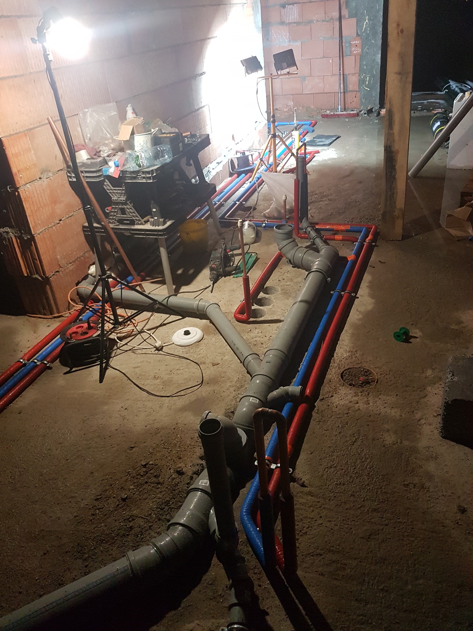 Instalacja wodno-kanalizacyjna w surowym stanie, rury szare i czerwono-niebieskie, oświetlenie budowlane, widok z góry na podłogę w trakcie montażu.