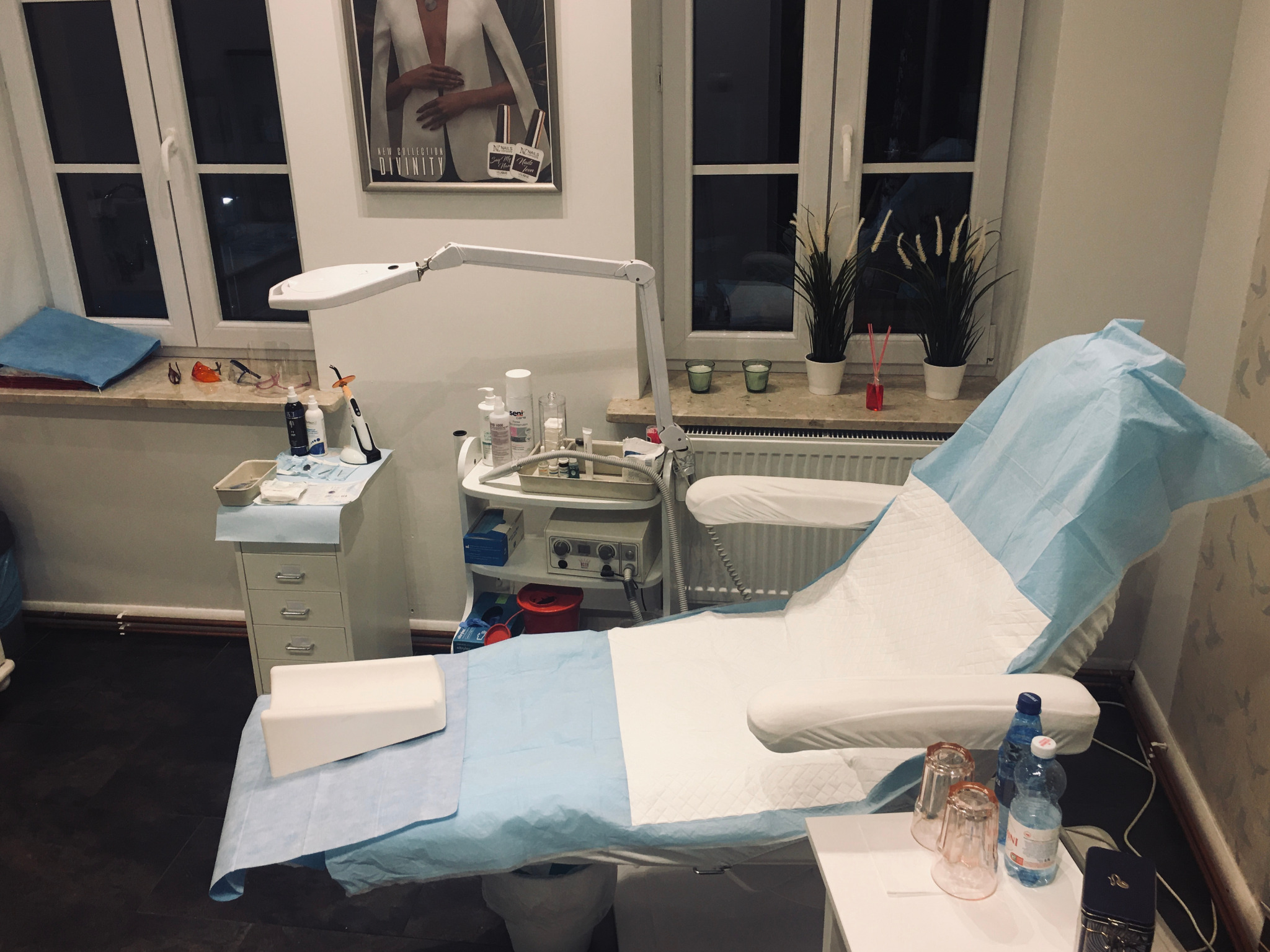 Kozetka kosmetyczna z niebieskim pokrowcem, lampa zabiegowa, szafka z akcesoriami kosmetycznymi i plakat z modelką w salonie kosmetycznym.