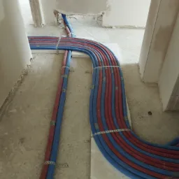 Układ czerwonych i niebieskich rur instalacji grzewczej/hydraulicznej ułożonych równolegle na podłodze w surowym stanie, zabezpieczone metalowymi klamrami.