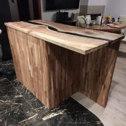 Wyspa kuchenna z blatem z drewna klejonego z żywicą epoksydową, widoczna na tle jadalni, z fragmentem osoby w lewym rogu. Podłoga z czarnego marmuru kontrastuje z jasnym drewnem.