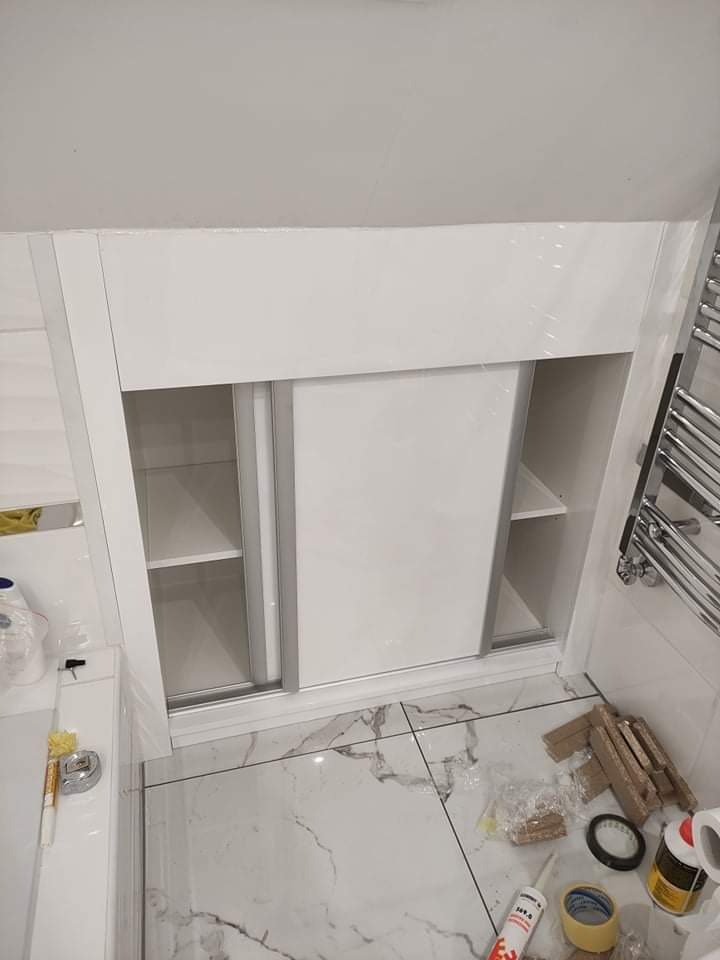 Biała szafka łazienkowa z przesuwnymi drzwiami w trakcie montażu, widoczne akcesoria montażowe i marmurowa posadzka.