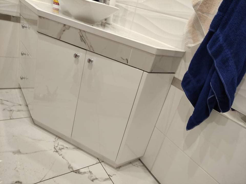 Biała szafka łazienkowa pod umywalkę z marmurowym blatem, fronty z chromowanymi uchwytami, widoczny fragment marmurowej podłogi i niebieski ręcznik.