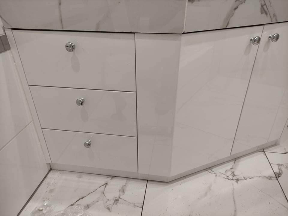 Biała, lakierowana szafka łazienkowa z trzema szufladami i szafką z drzwiczkami, chromowane gałki, marmurowy blat i podłoga z motywem marmuru.