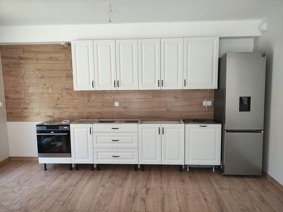 Wnętrze kuchni w trakcie montażu: białe szafki z czarnymi uchwytami, drewnopodobna ściana i podłoga, widoczna lodówka i piekarnik, brak blatu.
