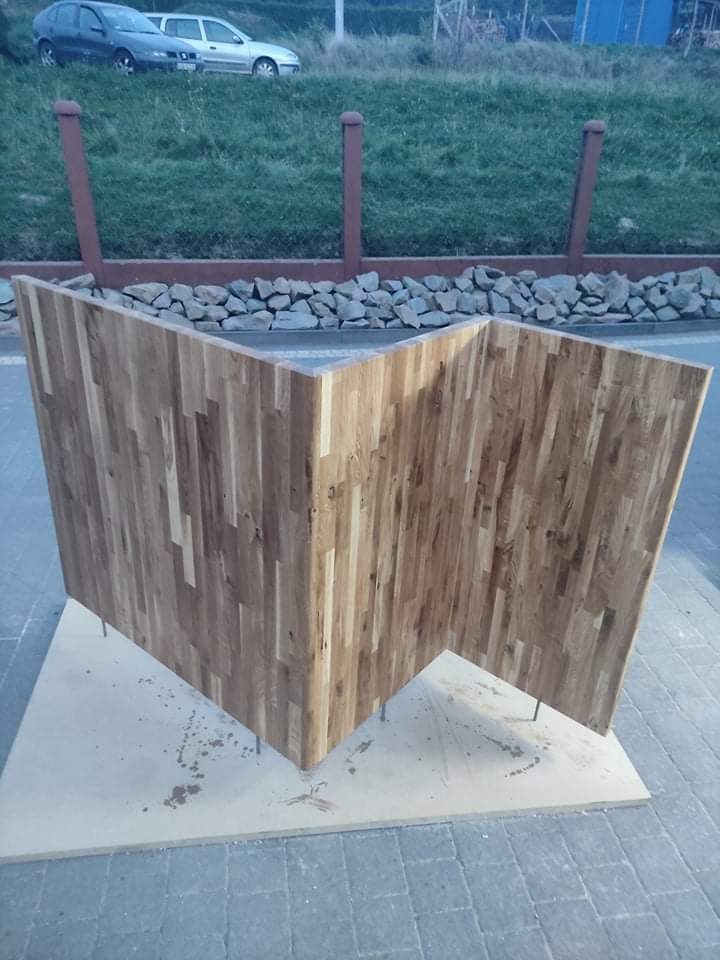 Surowy, trójkątny element z klejonych desek dębowych, ustawiony na płycie OSB na zewnątrz, widoczne metalowe pręty stabilizujące konstrukcję, w tle trawnik, kamienie i zaparkowane samochody.