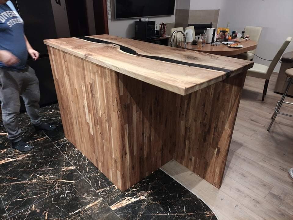 Wyspa kuchenna z blatem z drewna klejonego z żywicą epoksydową, widoczna na tle jadalni, z fragmentem osoby w lewym rogu. Podłoga z czarnego marmuru kontrastuje z jasnym drewnem.