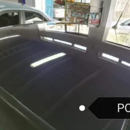 Błyszczący, ciemny lakier dachu samochodu po polerowaniu, odbijający światło lamp warsztatowych i okien budynku w tle.