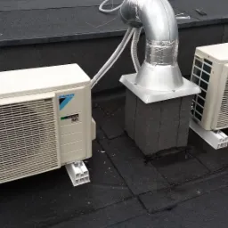 Dwa klimatyzatory Daikin zamontowane na płaskim, bitumicznym dachu, połączone przewodami z metalowym kominem wentylacyjnym. Na jednym z urządzeń leży śrubokręt.