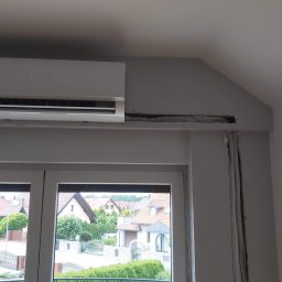 Maxcold Sp. z o.o. - Szary klimatyzator ścienny zamontowany nad oknem, widoczne ślady po instalacji przewodów w ścianie, w tle widok na osiedle domów jednorodzinnych.