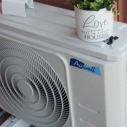 Maxcold Sp. z o.o. - Biała zewnętrzna jednostka klimatyzacji Airwell z doniczką z napisem 'Love grows best in little houses' stojącą na górze, widoczne mocowanie na boku.