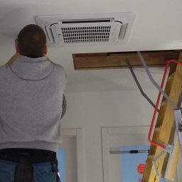 Maxcold Sp. z o.o. - Pracownik na drabinie montuje kasetonową jednostkę klimatyzacyjną w suficie podwieszanym, widoczny otwór instalacyjny i narzędzia w kieszeniach.