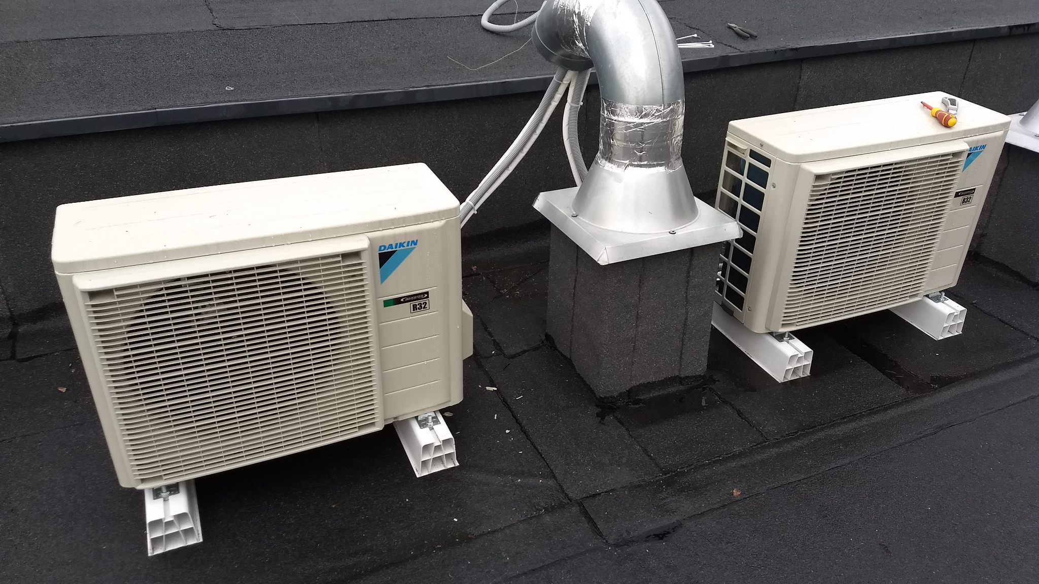 Dwa klimatyzatory Daikin zamontowane na płaskim, bitumicznym dachu, połączone przewodami z metalowym kominem wentylacyjnym. Na jednym z urządzeń leży śrubokręt.