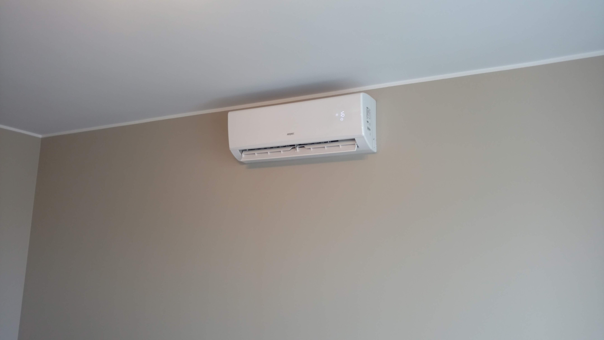 Biała jednostka klimatyzacji zamontowana na ścianie w jasnym pomieszczeniu, widoczny wyświetlacz z temperaturą.