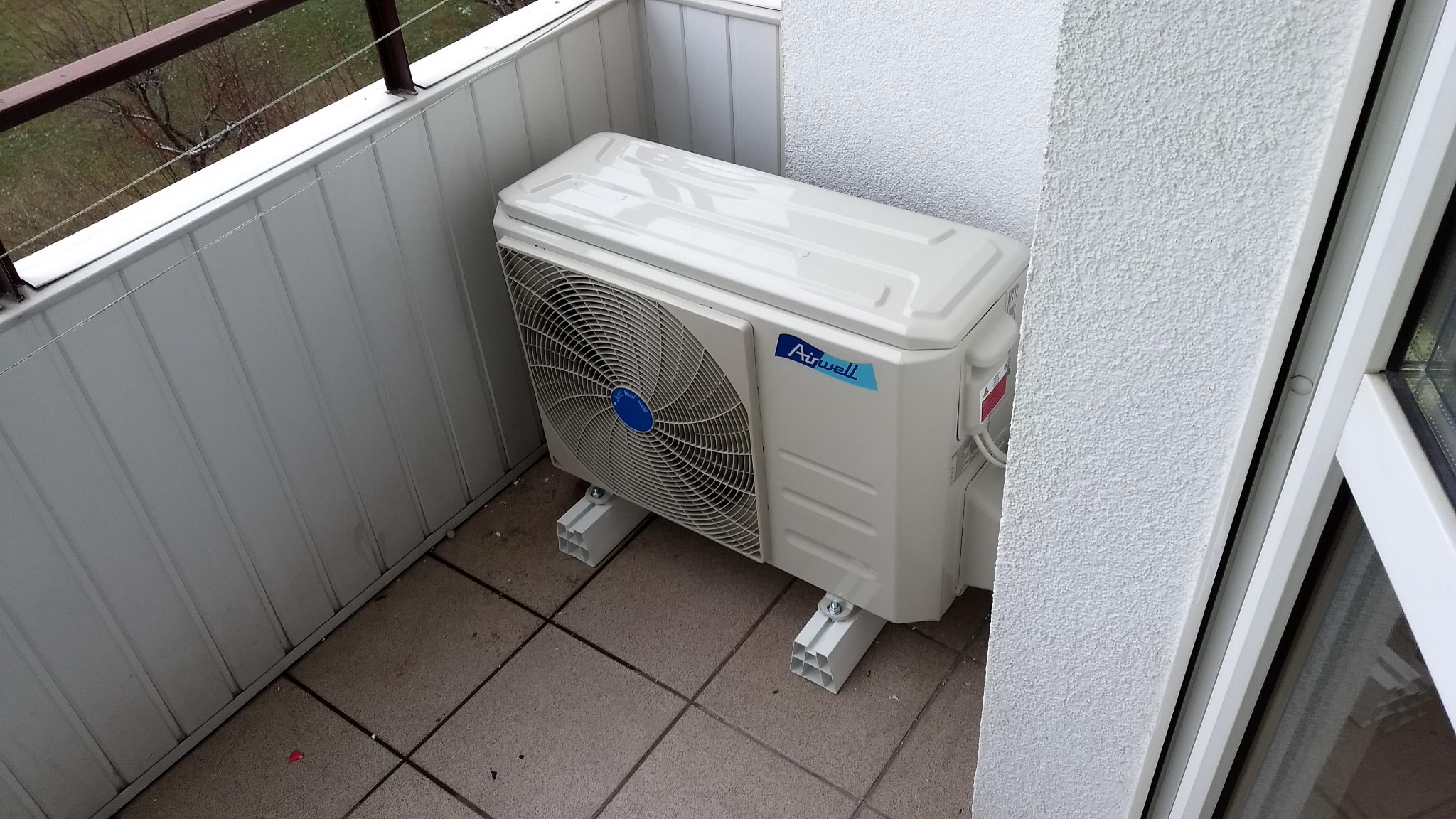 Zewnętrzna jednostka klimatyzacji marki Airwell zamontowana na balkonie, widoczne wsporniki montażowe i płytki balkonowe.