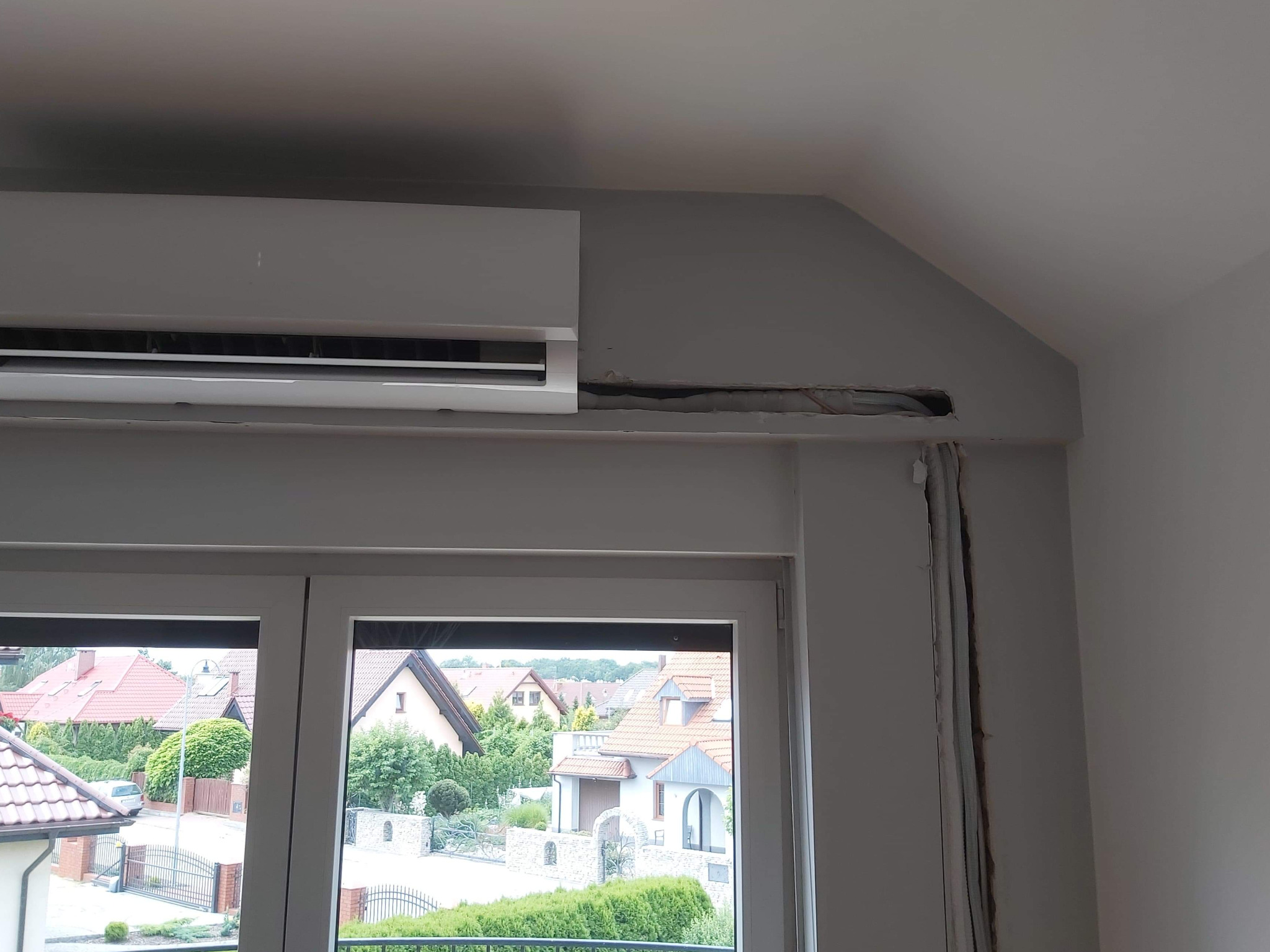 Szary klimatyzator ścienny zamontowany nad oknem, widoczne ślady po instalacji przewodów w ścianie, w tle widok na osiedle domów jednorodzinnych.