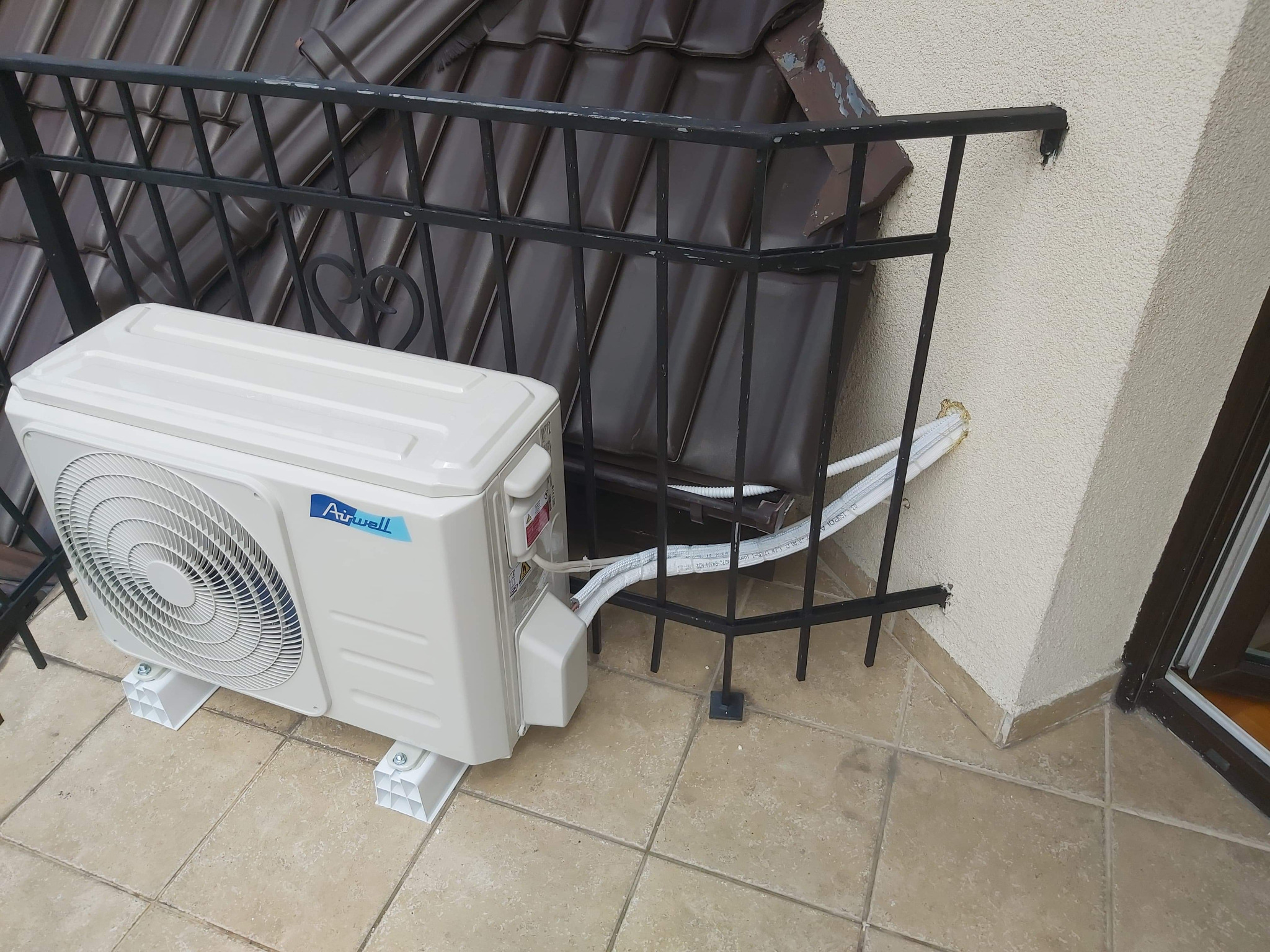 Biała jednostka zewnętrzna klimatyzatora marki Airwell zamontowana na balkonie z ciemną balustradą i brązowymi dachówkami w tle.