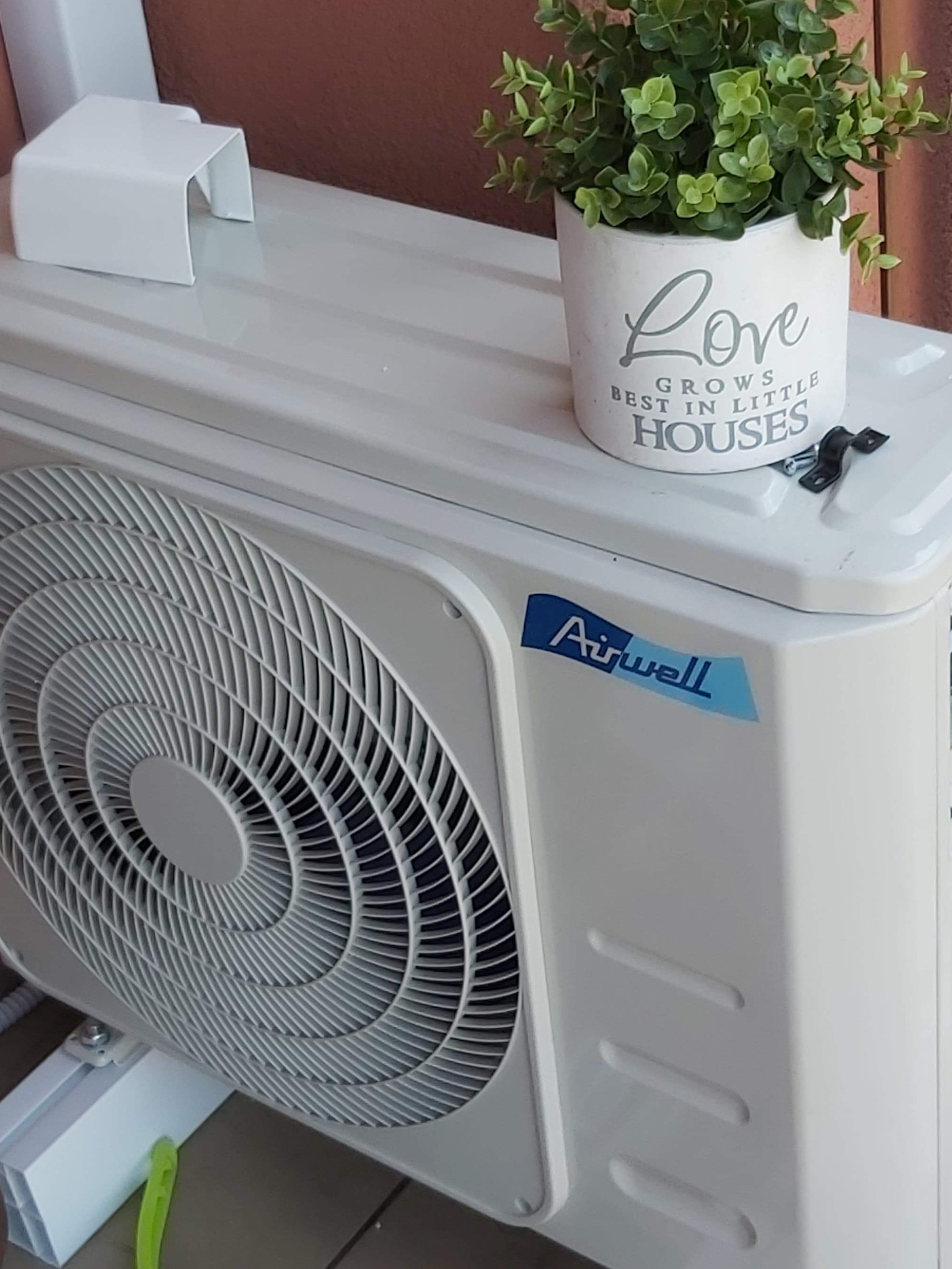 Biała zewnętrzna jednostka klimatyzacji Airwell z doniczką z napisem 'Love grows best in little houses' stojącą na górze, widoczne mocowanie na boku.