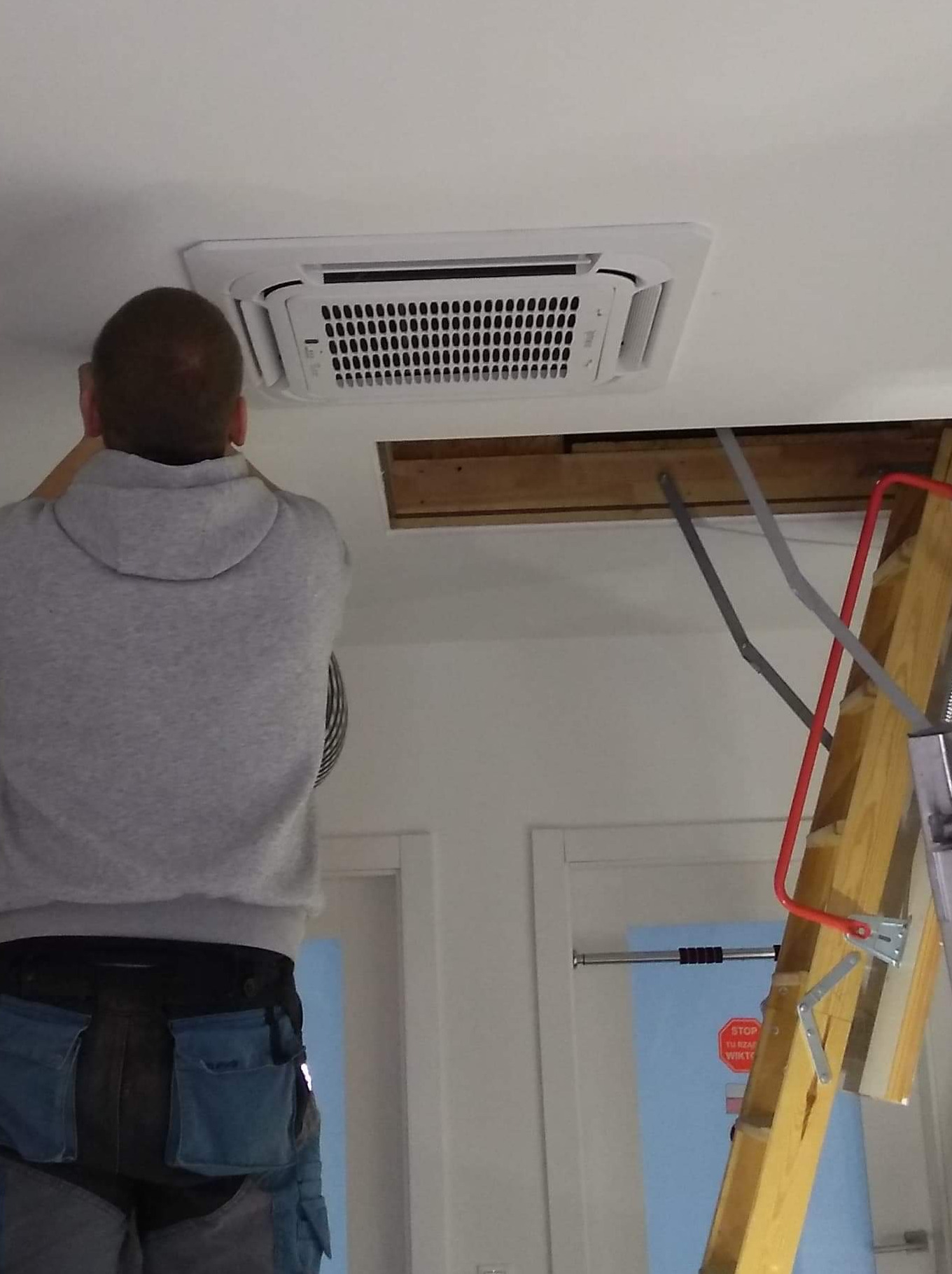 Pracownik na drabinie montuje kasetonową jednostkę klimatyzacyjną w suficie podwieszanym, widoczny otwór instalacyjny i narzędzia w kieszeniach.