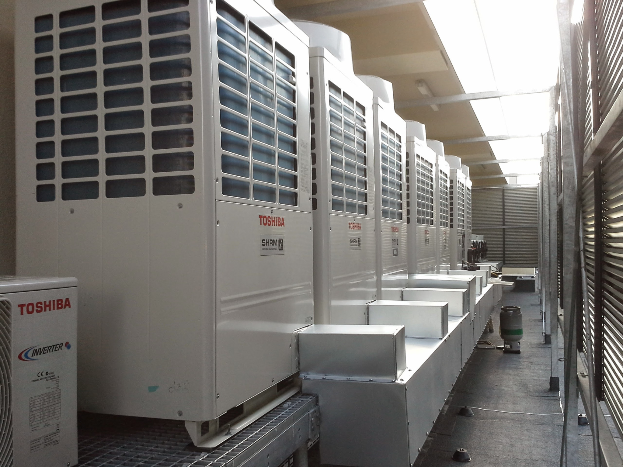 Rząd białych jednostek zewnętrznych klimatyzacji Toshiba SHRM-e ustawionych na dachu budynku komercyjnego, widok perspektywiczny z boku, z widocznymi kratkami wentylacyjnymi i elementami instalacji.