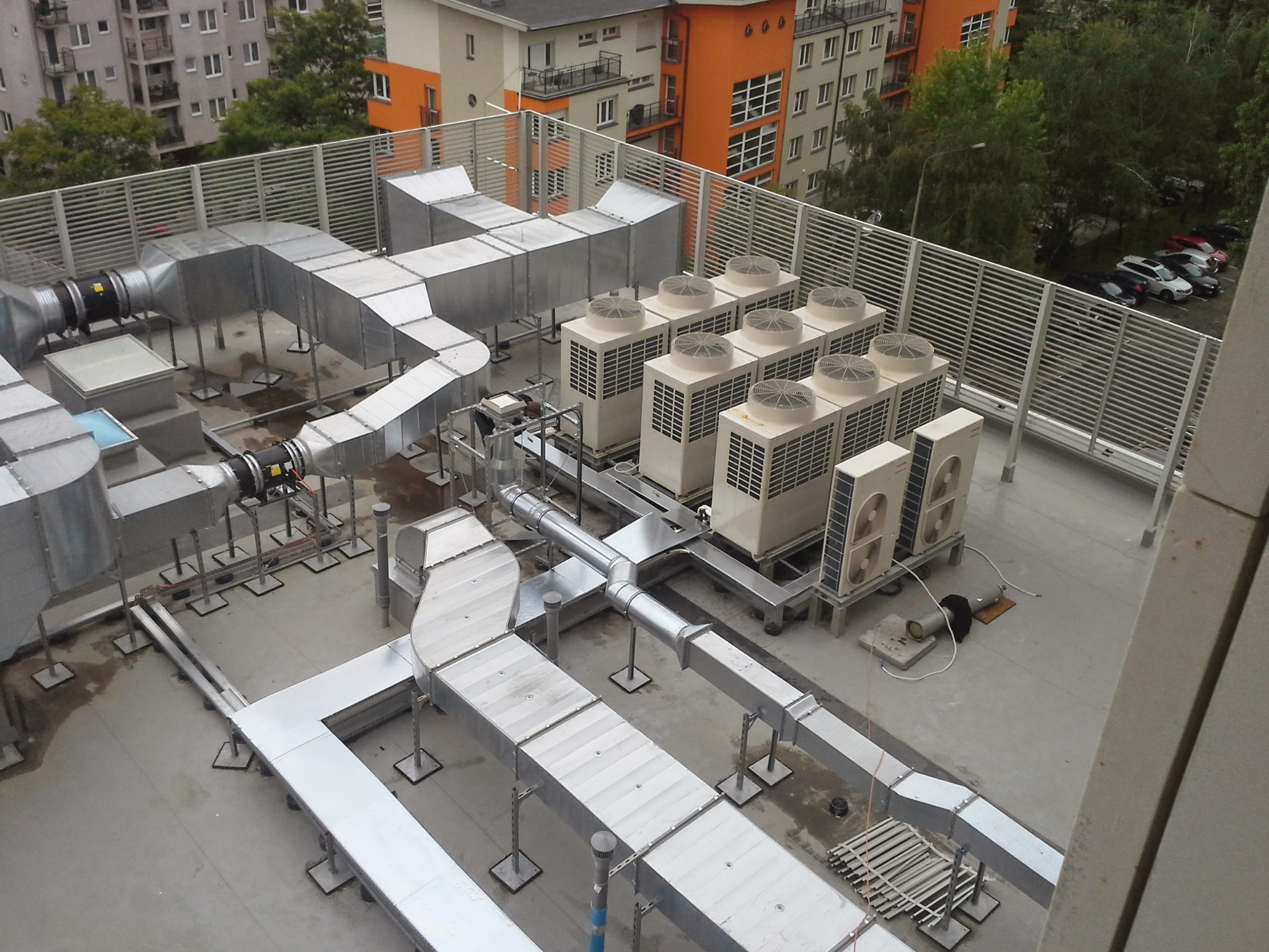 Widok z góry na rozbudowany system wentylacyjny z kanałami i centralami na dachu budynku, zabezpieczony ażurową balustradą.