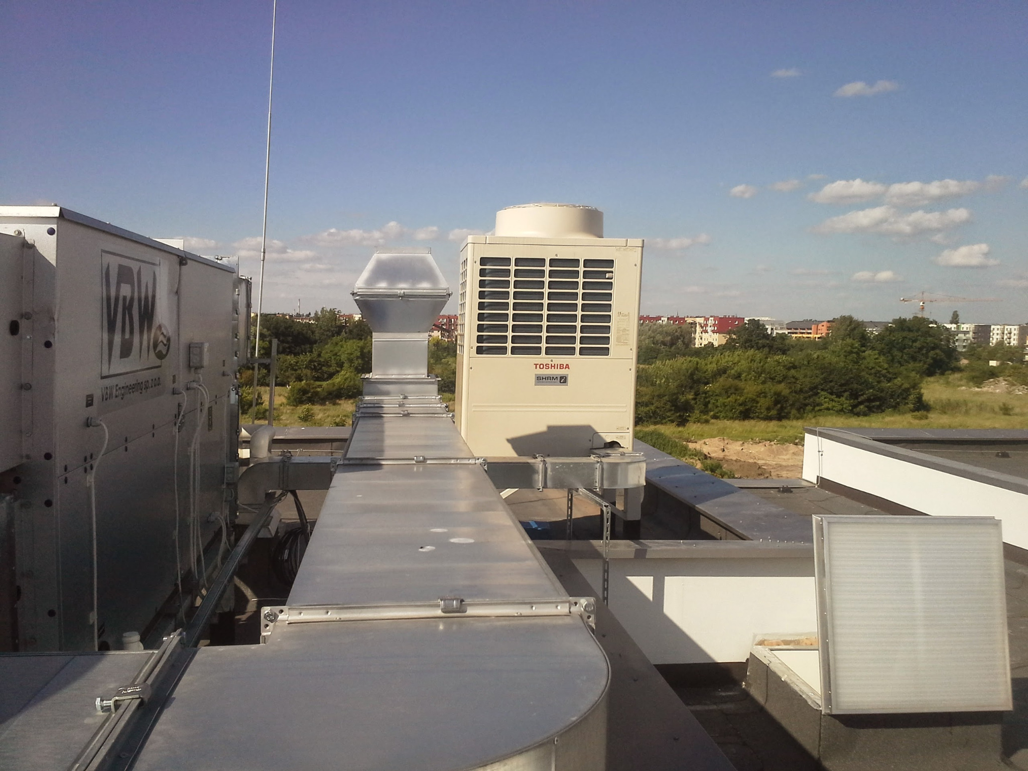 Instalacja wentylacyjna na dachu budynku we Wrocławiu, widoczne kanały wentylacyjne, centrala wentylacyjna marki Toshiba i elementy systemu rekuperacji. W tle zabudowa miejska i błękitne niebo.
