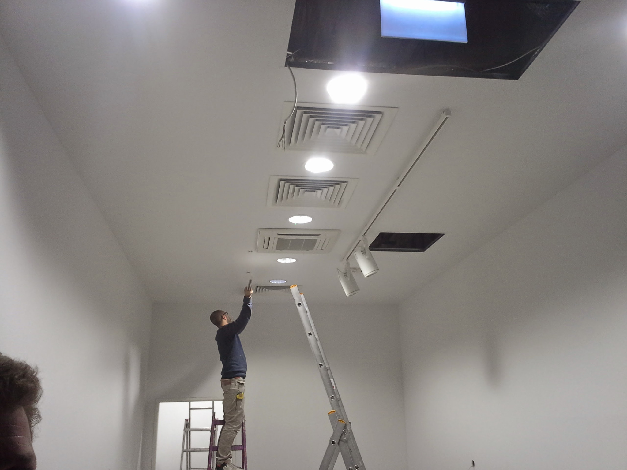 Pracownik na drabinie montuje kratkę wentylacyjną w suficie podwieszanym w pomieszczeniu komercyjnym z oświetleniem szynowym.