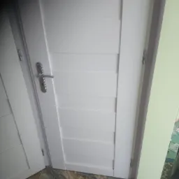 Białe drzwi wewnętrzne z poziomymi panelami, widoczny srebrny klamek i fragment podłogi z wzorem marmuru.