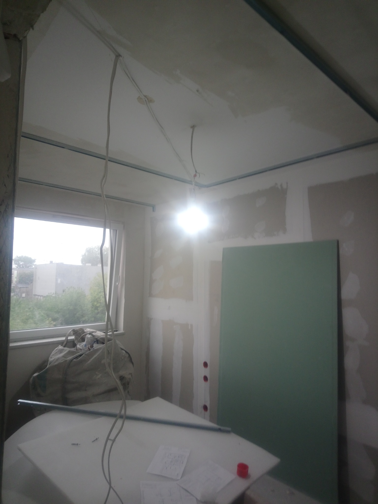 Wnętrze w trakcie prac: widoczne profile pod sufitem, przewody elektryczne zwisające z sufitu, częściowo położona gładź na ścianach, płyta gipsowo-kartonowa oparta o ścianę, okno z widokiem...