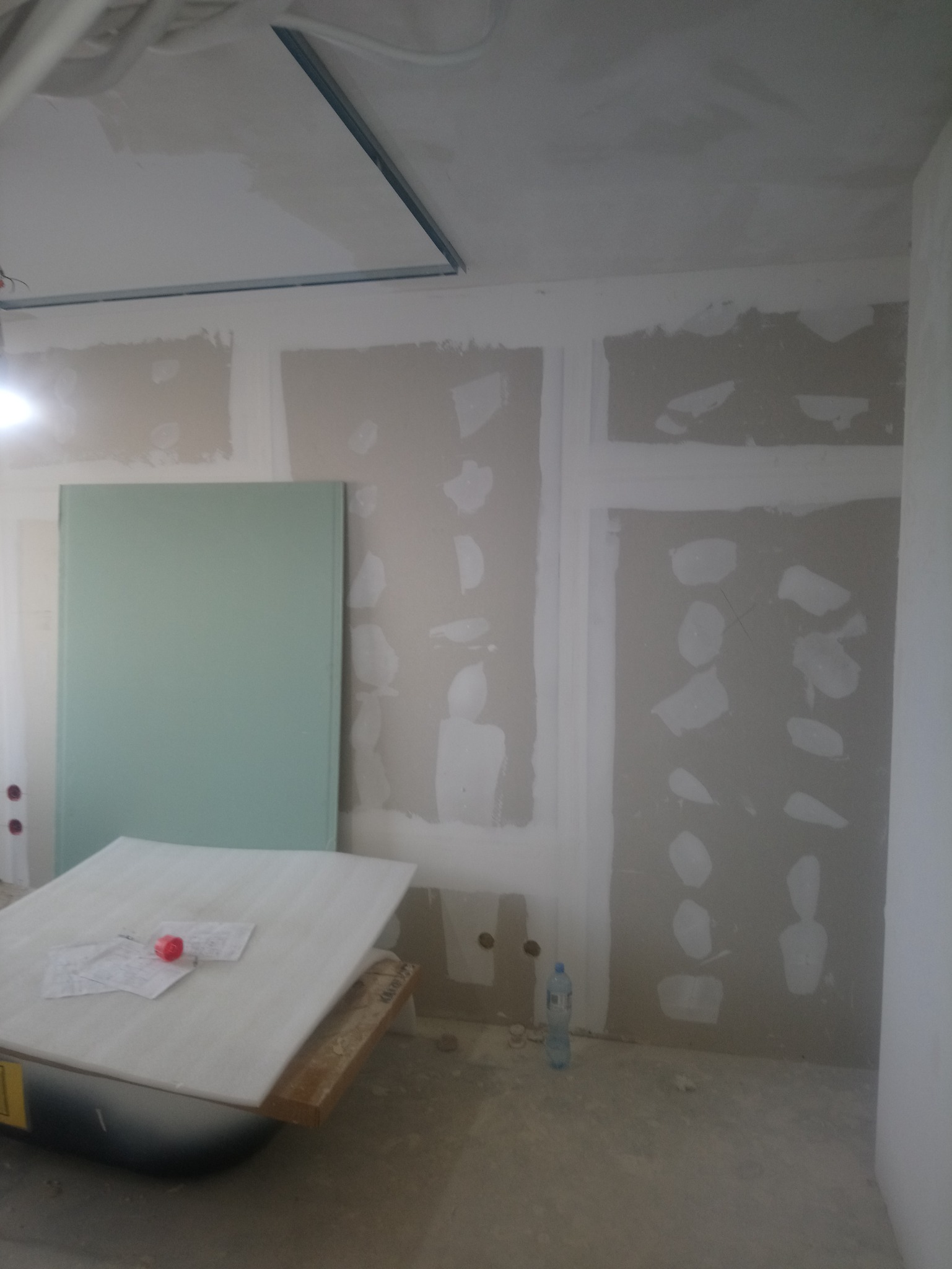 Wnętrze w trakcie prac wykończeniowych, widoczne płyty kartonowo-gipsowe na ścianach, szpachlowane łączenia, profil sufitowy, płyta gipsowa oparta o ścianę, butelka wody na podłodze.