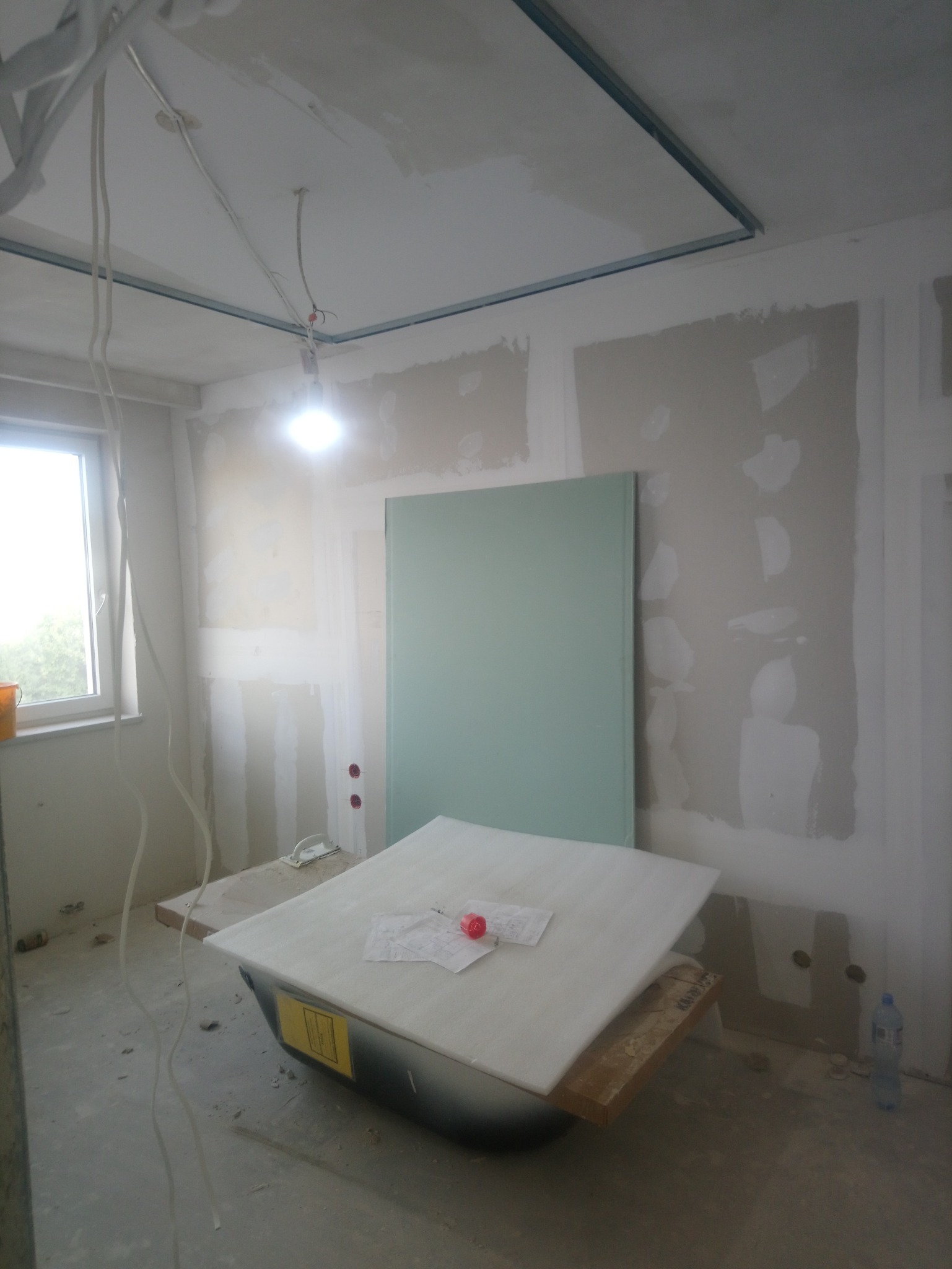 Pokój w trakcie prac wykończeniowych: widoczne płyty kartonowo-gipsowe na ścianach, metalowa konstrukcja sufitu podwieszanego, tymczasowe oświetlenie, wanna wykorzystywana jako podstawa do prac...