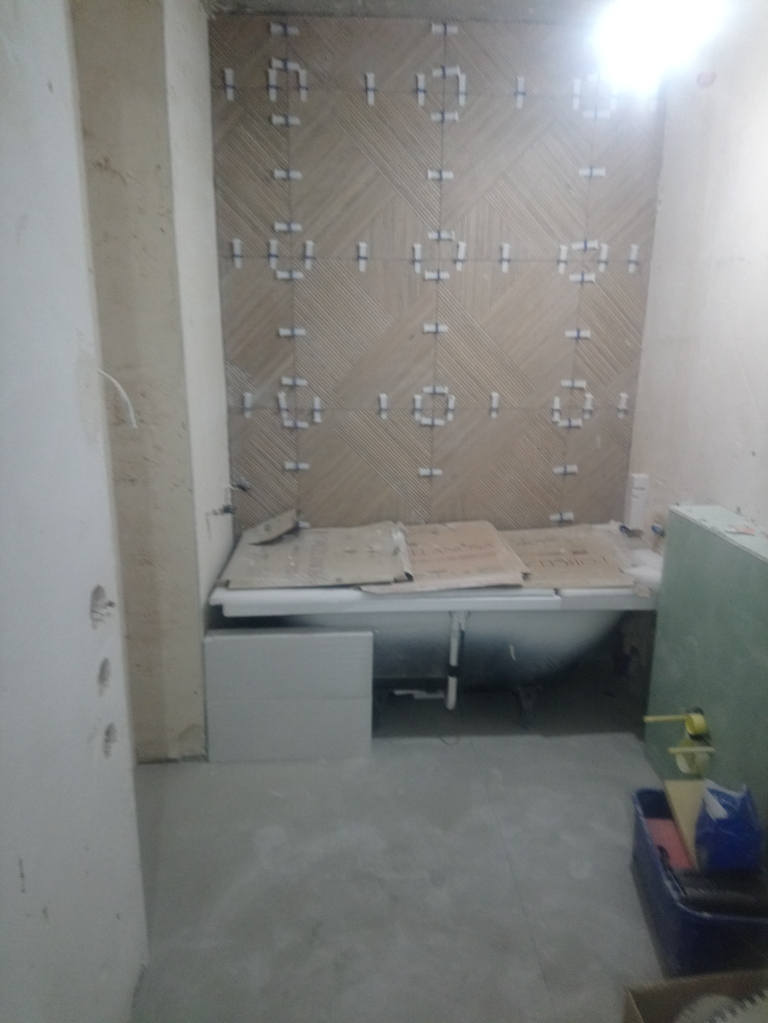 Łazienka w trakcie prac wykończeniowych: świeżo położona beżowa glazura na ścianie z użyciem krzyżyków dystansowych, wanna zabezpieczona kartonem, widoczne narzędzia i materiały budowlane.