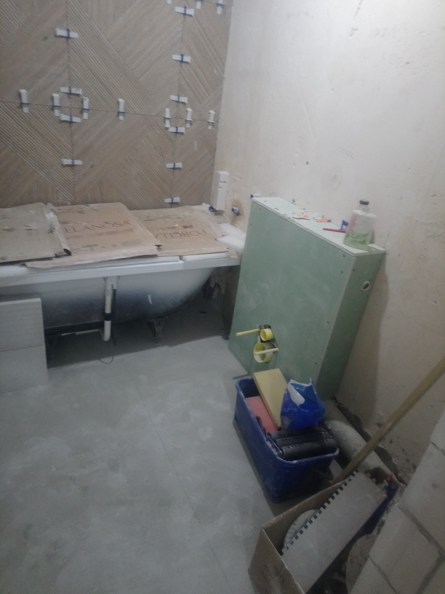 Łazienka w trakcie remontu: częściowo wykończona ściana z płytek ceramicznych z krzyżykami dystansowymi, obudowana wanna z kartonami ochronnymi, płyta karton gips z wystającymi rurami, niebieski...