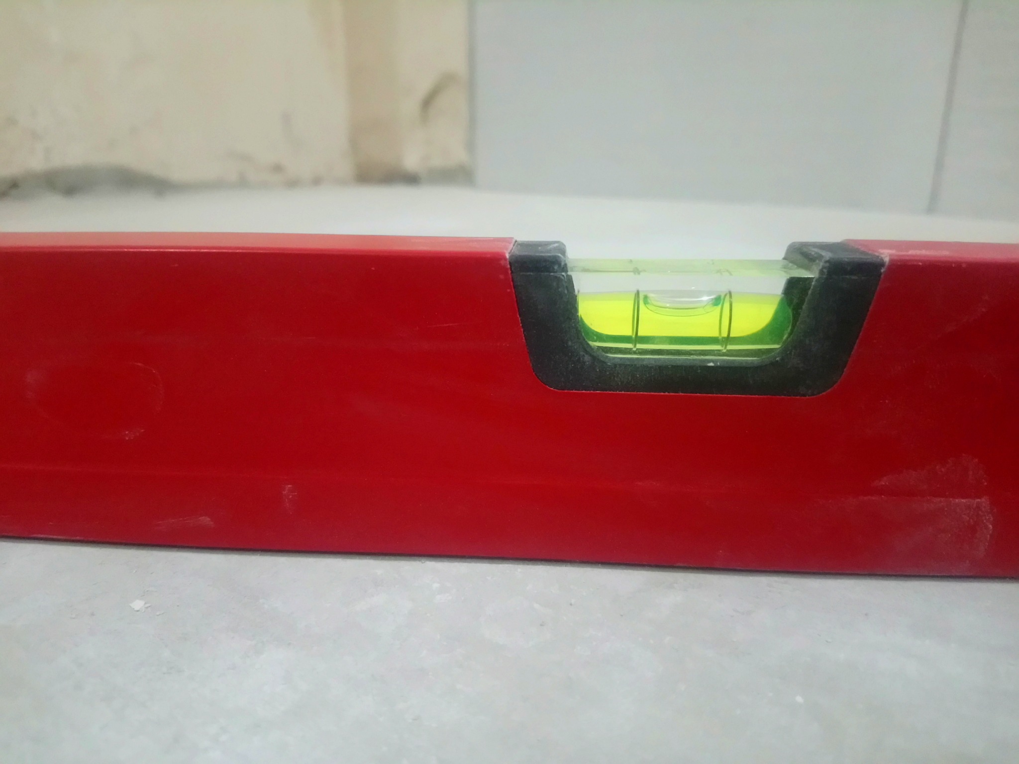 Czerwona poziomica na szarej, prawdopodobnie betonowej, powierzchni w trakcie prac wykończeniowych; widoczny fragment ściany w tle.