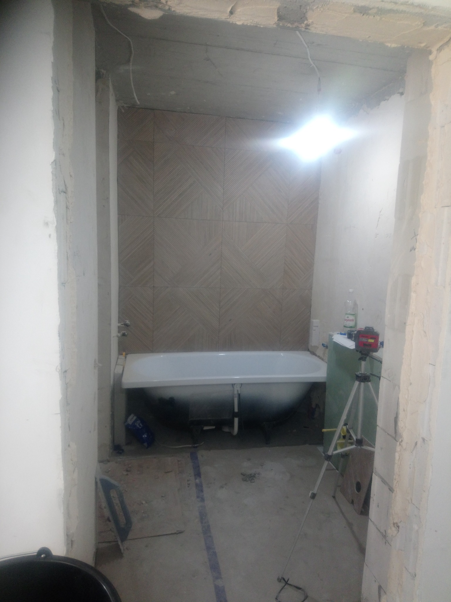 Łazienka w trakcie remontu: świeżo ułożona geometryczna glazura na ścianie za białą wanną, widoczny laser krzyżowy na statywie, surowy beton i ślady zaprawy na ścianach.