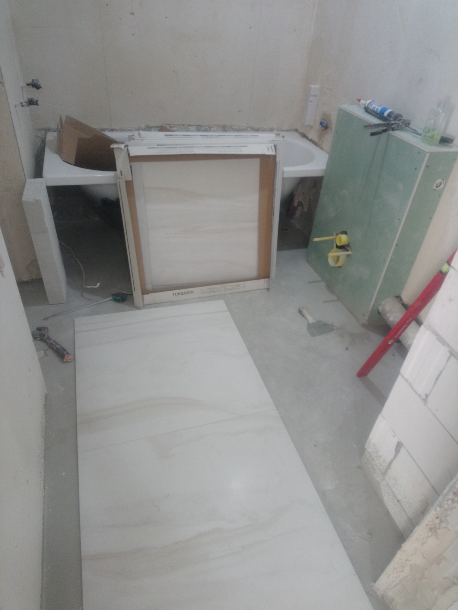 Przygotowane do montażu płytki podłogowe o jasnym, marmurkowym wzorze, obok wanna w trakcie obudowy, widoczne narzędzia glazurnicze i płyta gipsowo-kartonowa.