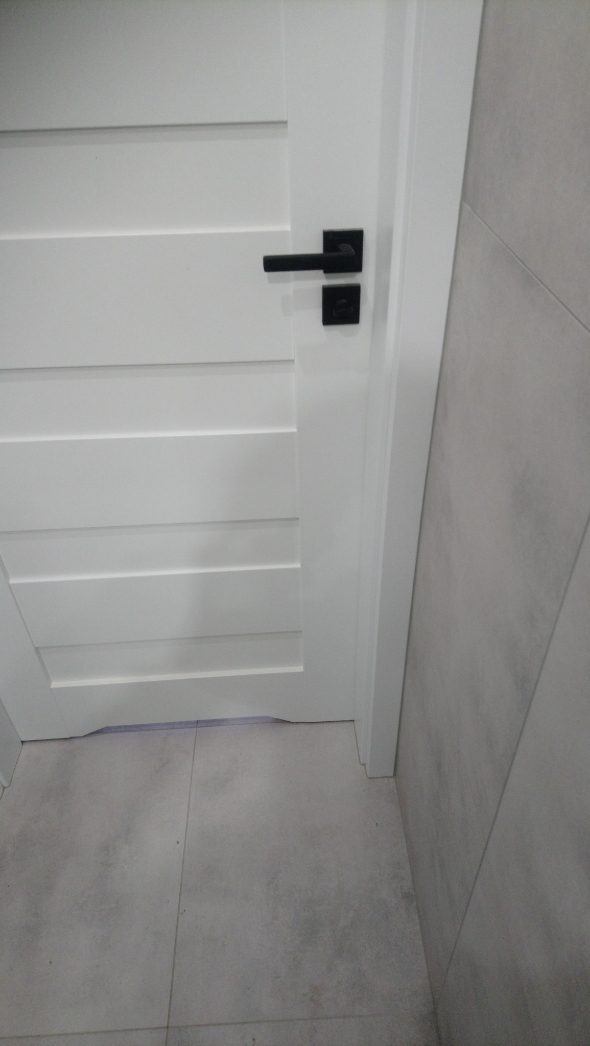 Białe drzwi wewnętrzne z poziomymi panelami i czarną, kwadratową klamką, widoczne na tle szarej płytki podłogowej i ściennej.