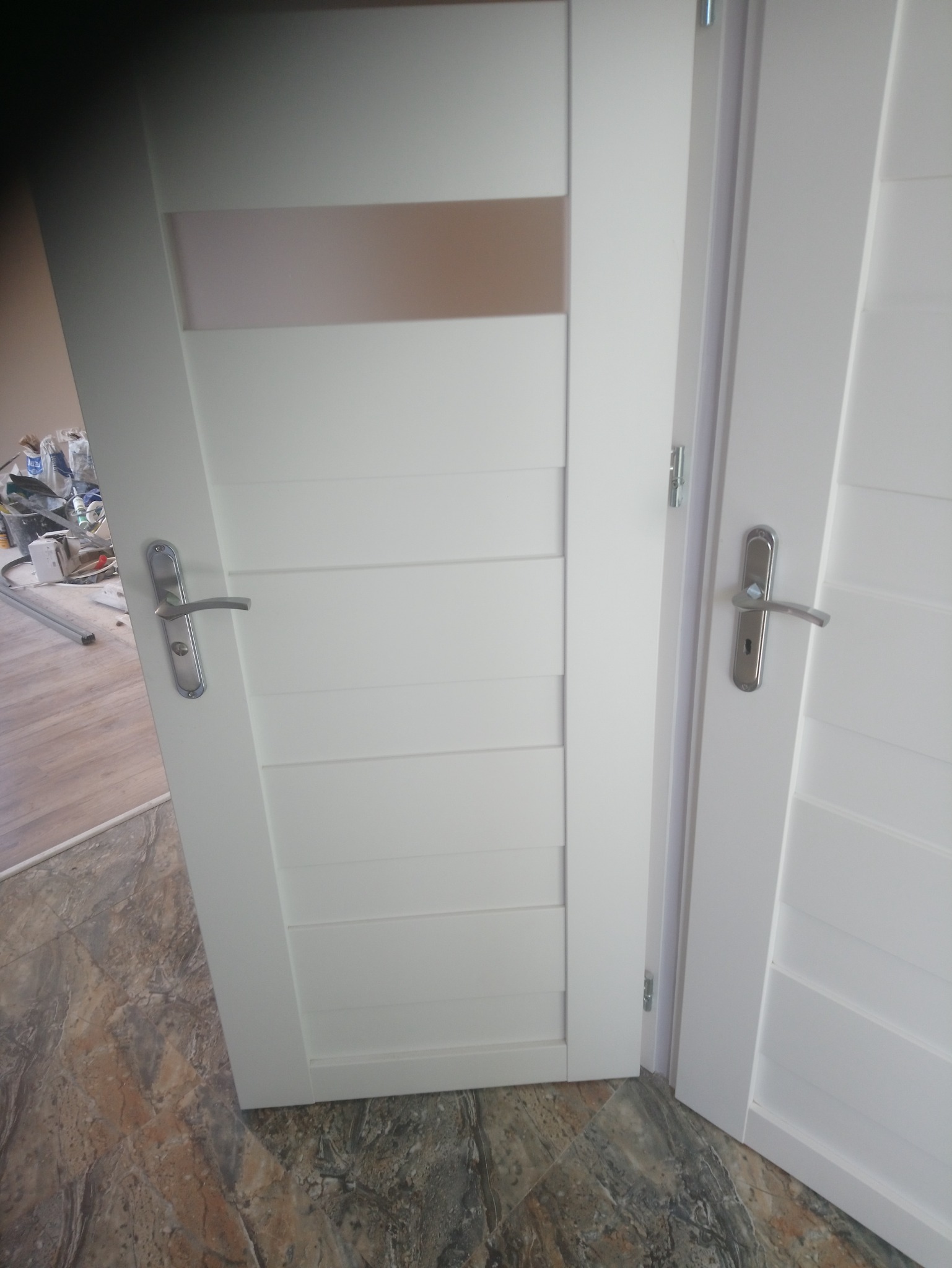 Białe, częściowo otwarte drzwi wewnętrzne z poziomymi wgłębieniami i szklanym panelem u góry, widoczne narzędzia i materiały budowlane w tle.