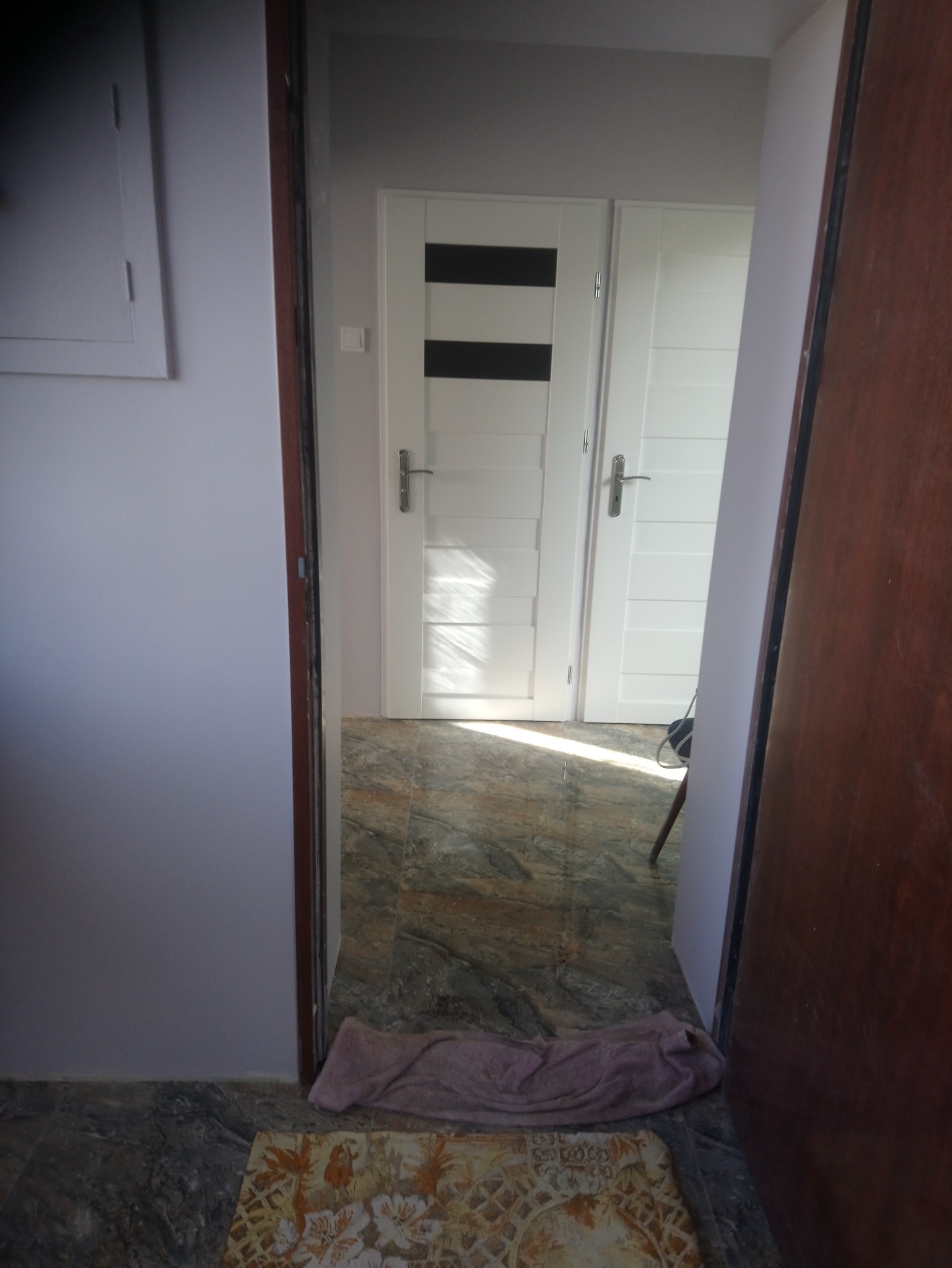 Widok na dwie białe, nowoczesne drzwi wewnętrzne z czarnymi poziomymi paskami, widziane zza otwartych, ciemnych drzwi. Na podłodze leży dywanik i zwinięty ręcznik.