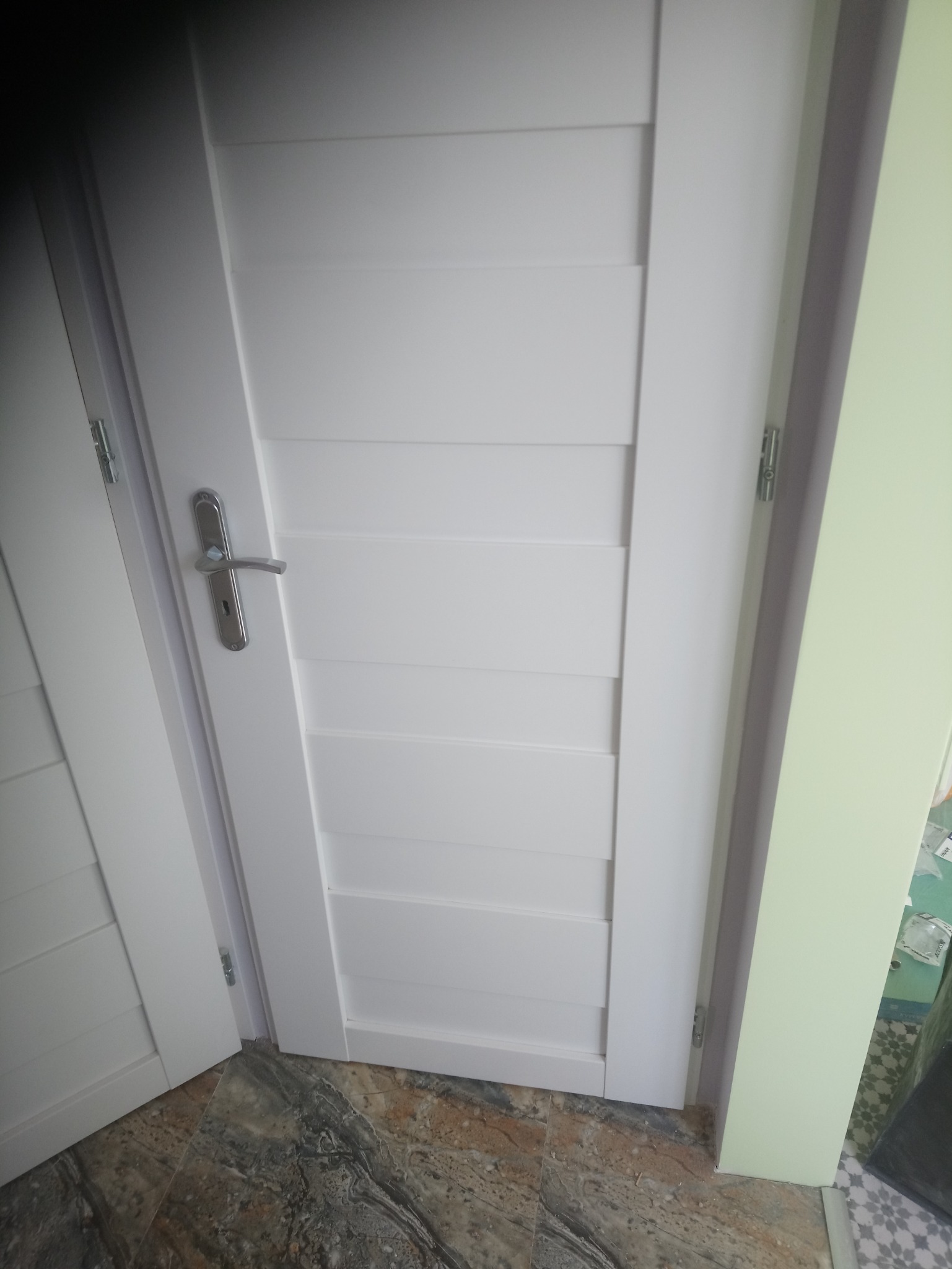 Białe drzwi wewnętrzne z poziomymi panelami, widoczny srebrny klamek i fragment podłogi z wzorem marmuru.