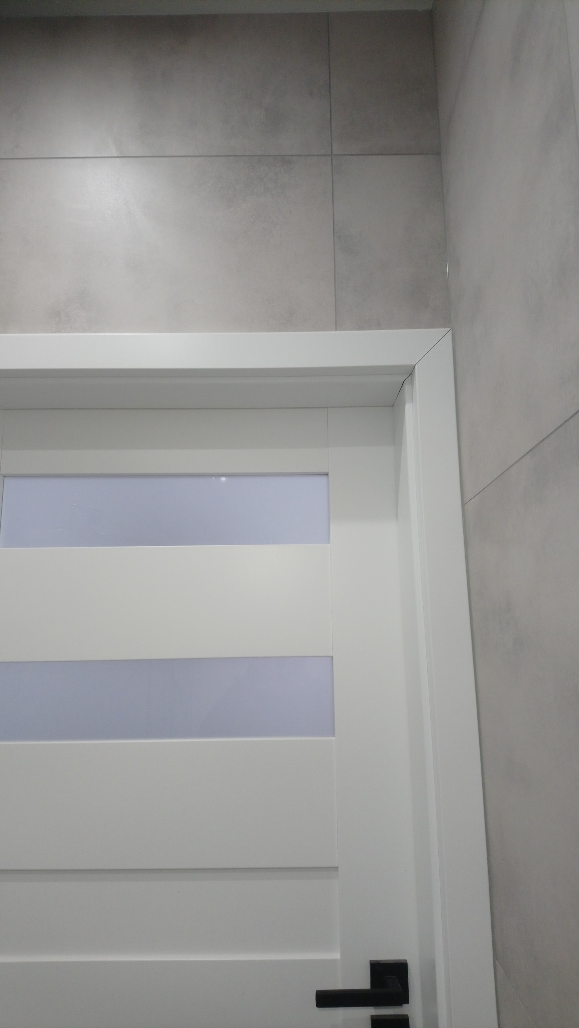 Nowoczesne, białe drzwi z przeszkleniami, zamontowane w łazience z szarymi, matowymi płytkami. Widoczna czarna klamka.