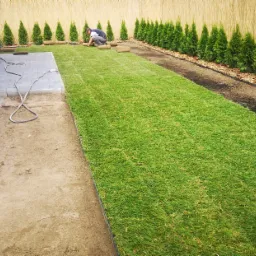 Układanie zielonej murawy z rolki w ogrodzie, widoczny pracownik w trakcie pracy, fragment nawodnienia, rząd tuj przy płocie z bambusa.