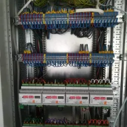 Rozdzielnica elektryczna z licznymi przewodami w różnych kolorach połączonymi z listwami zaciskowymi i modułami, widoczne oznaczenia na urządzeniach.