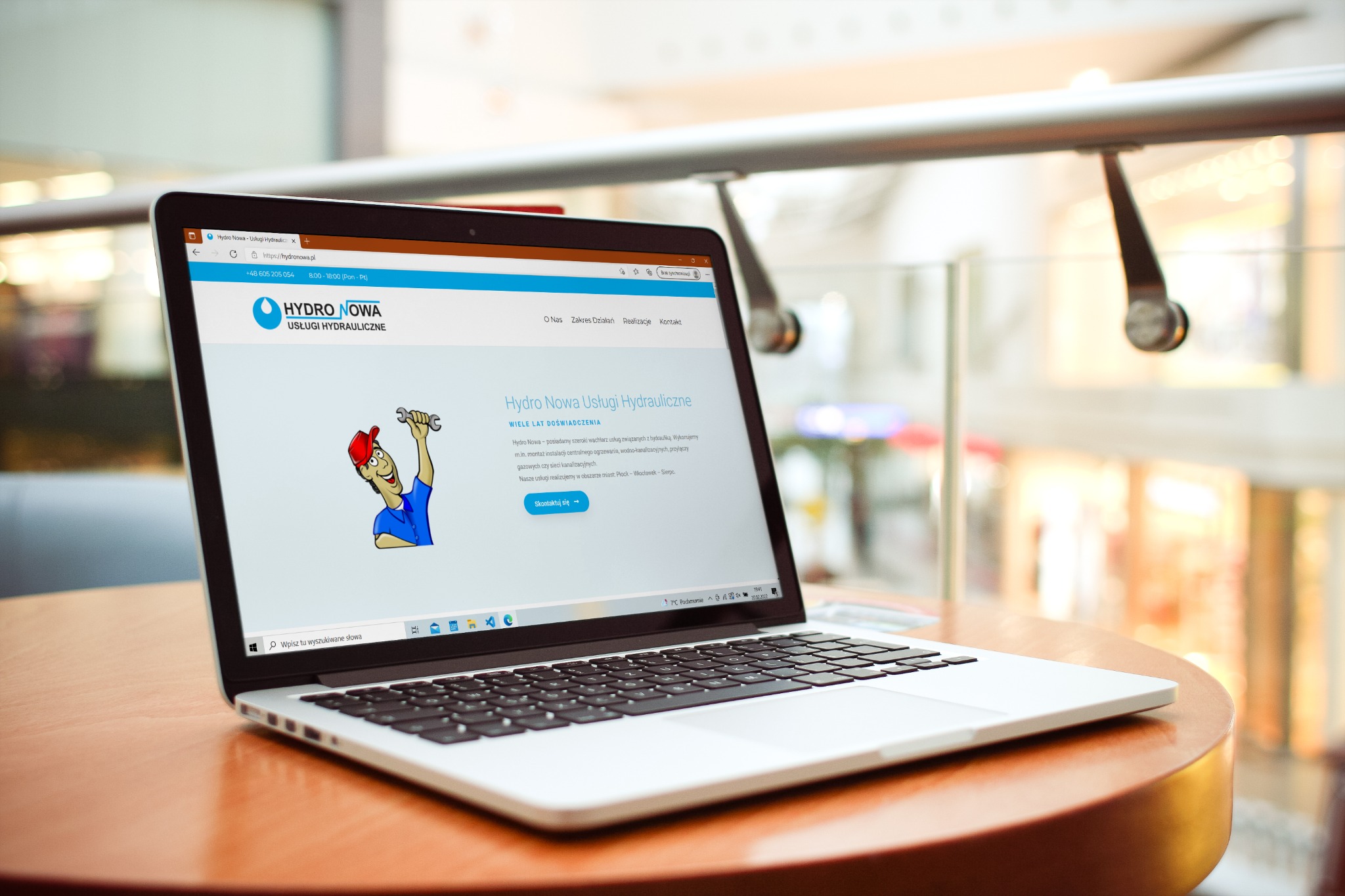 Laptop wyświetla stronę internetową firmy hydraulicznej z Torunia, www.hydronowa.pl, prezentującą usługi i dane kontaktowe, umieszczony na okrągłym stole w przestrzeni publicznej.