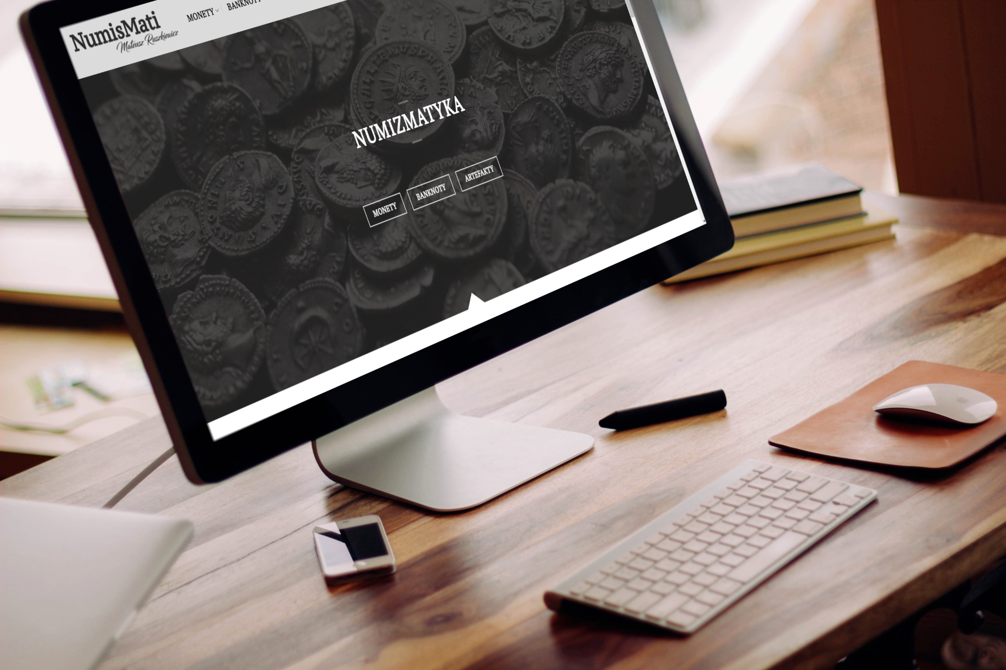 Monitor wyświetla stronę internetową sklepu numizmatycznego z motywem monet, leżący na drewnianym biurku obok klawiatury, myszki, smartfona i pióra.