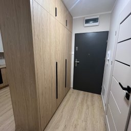 Jasna szafa wnękowa z długimi, czarnymi uchwytami, dopasowana do koloru podłogi. Obok drzwi wejściowe i wewnętrzne. Minimalistyczny design.