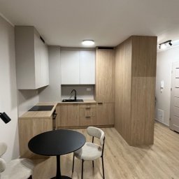 Jasna kuchnia z drewnianymi frontami i białymi szafkami wiszącymi. Na pierwszym planie czarny stół z krzesłami. Nowoczesny design, minimalistyczne wykończenie.