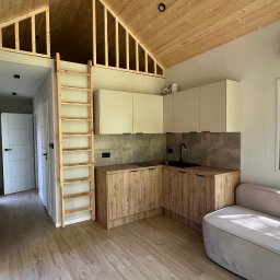 Drewniana drabina prowadzi na antresolę w jasnej kuchni z drewnianymi szafkami i szarą ścianą. W tle widoczny fragment sofy i okno z widokiem na zieleń.