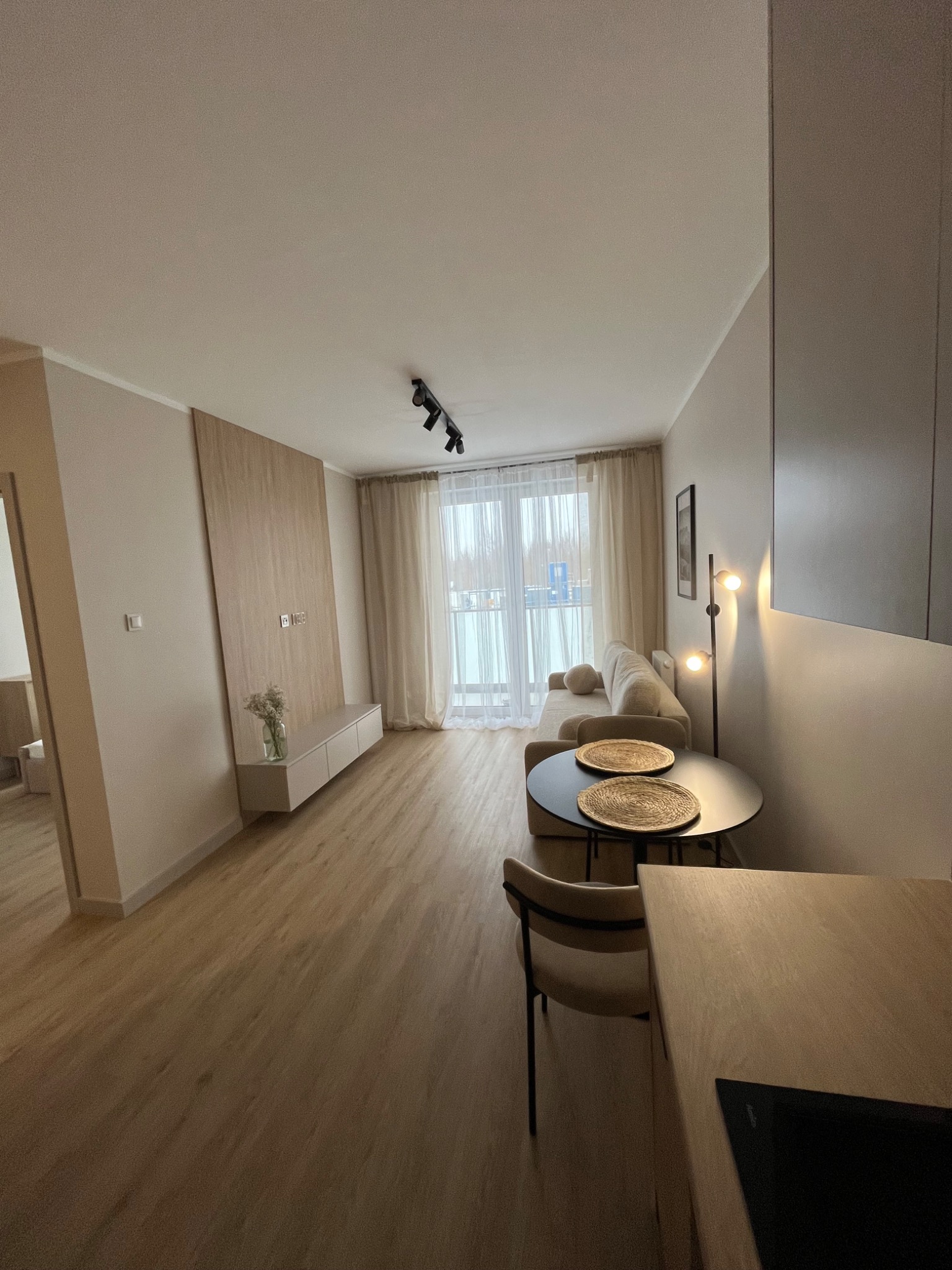 Jasne wnętrze mieszkania z drewnianą podłogą, beżową sofą i czarnym stolikiem. Na ścianie drewniana zabudowa TV z szafką. Widok na balkon przez okno z zasłonami.