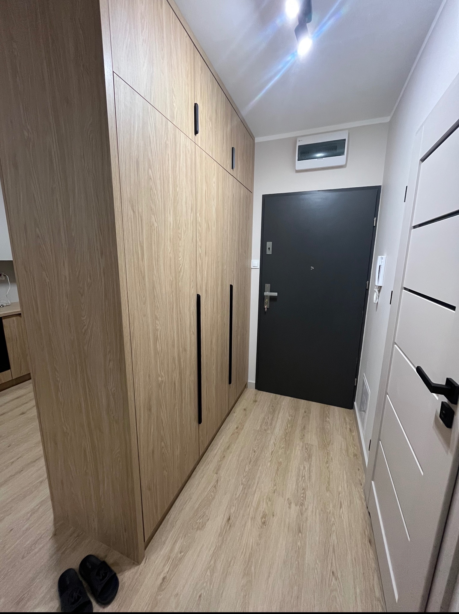 Jasna szafa wnękowa z długimi, czarnymi uchwytami, dopasowana do koloru podłogi. Obok drzwi wejściowe i wewnętrzne. Minimalistyczny design.